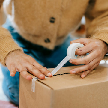 Person taping a cardboard box with a roll of tape, wearing a brown sweater and blue pants.